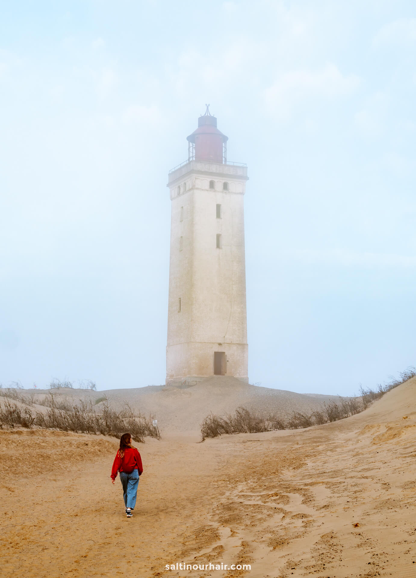 A woman in a red jacket walks toward a tall, beige lighthouse called Rubjerg Knude surrounded by sand and sparse vegetation on a foggy day—a classic scene for any Denmark itinerary.
