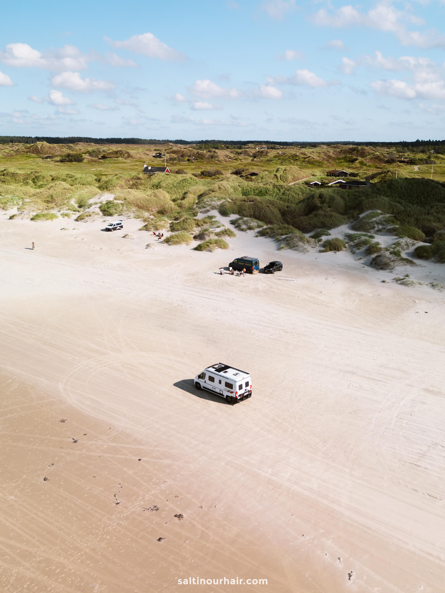 A white camper van drives on a wide sandy beach is the perfect addition to any Denmark road trip itinerary, with several vehicles and grassy dunes in the background under a partly cloudy sky.