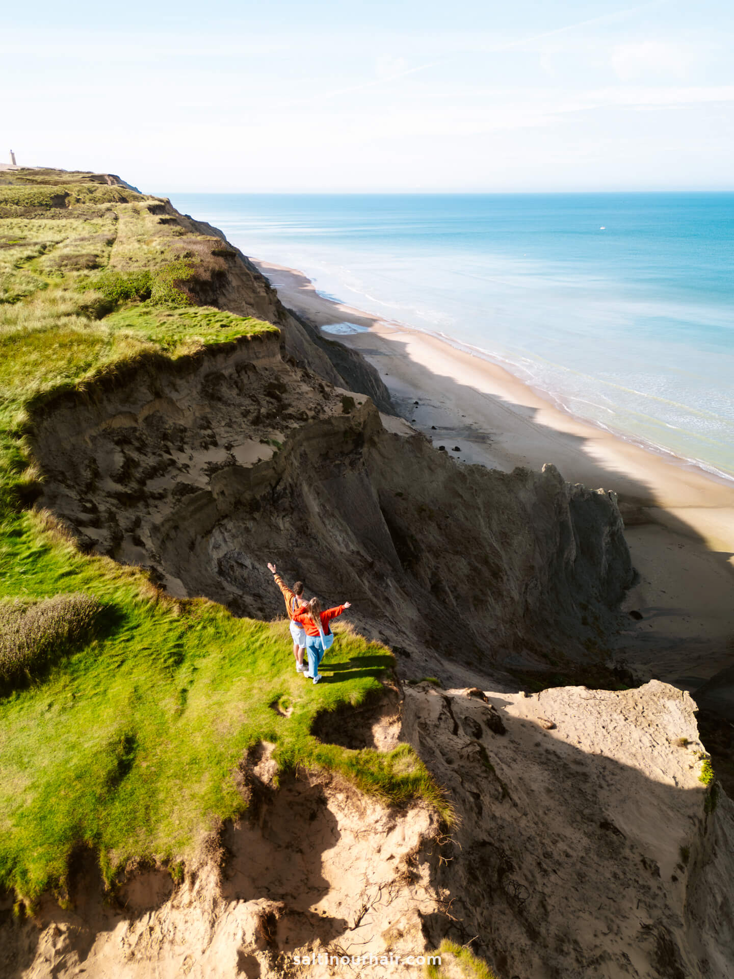 A person stands near the edge of a grassy cliff, perhaps following their Denmark itinerary, gazing over a beach and calm blue sea beneath a clear sky.