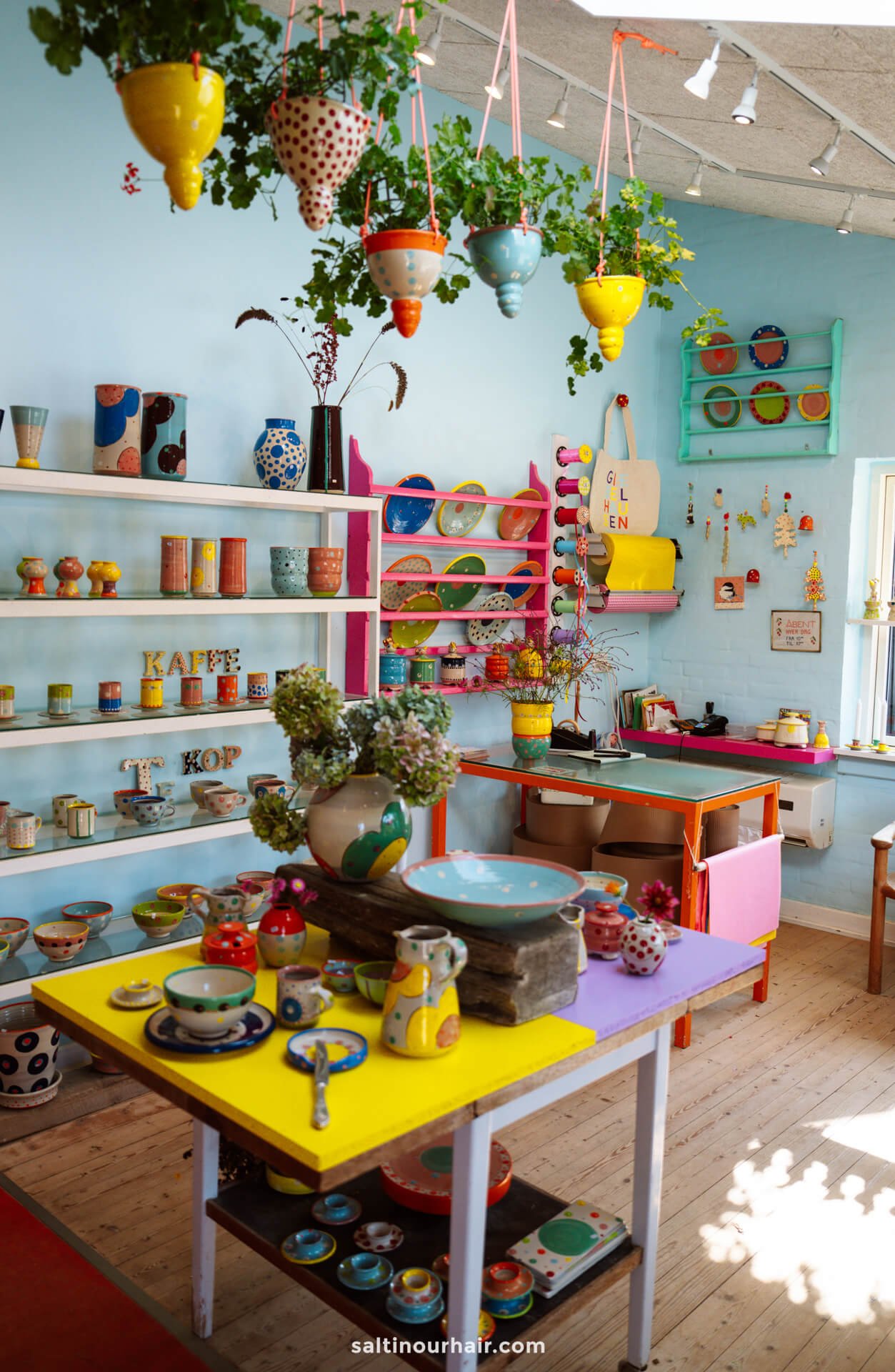 A brightly colored pottery shop in Lønstrup, perfect for any Denmark itinerary, displays various handmade ceramics—mugs, bowls, and vases—along with hanging plants and shelves against pastel blue walls.
