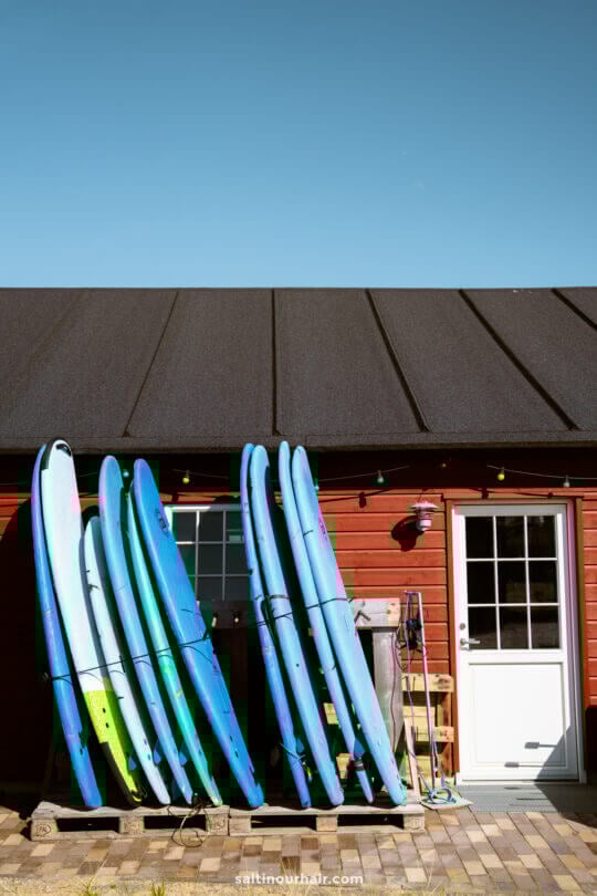 Several surfboards are propped against a red wooden building with a black roof, next to a white door and outdoor string lights—an inviting spot for any Denmark road trip itinerary under a clear blue sky.