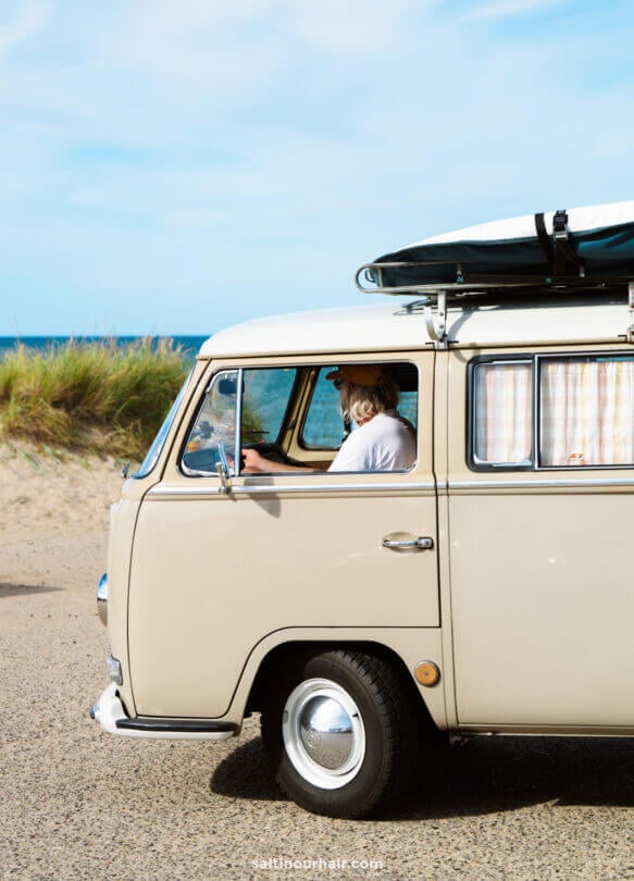 A person drives a beige vintage van with a roof rack near a sandy beach and grassy dunes under a blue sky, capturing the spirit of a classic Denmark road trip.