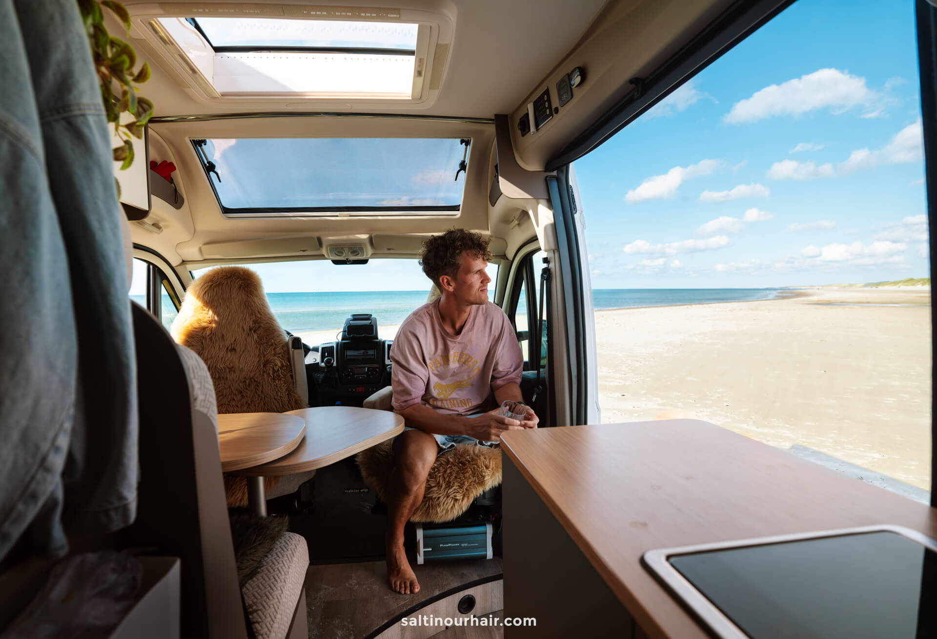A man sits inside a camper van parked by a sandy beach, looking out at the ocean through the open side door under a partly cloudy sky—an idyllic stop on his Denmark road trip itinerary.