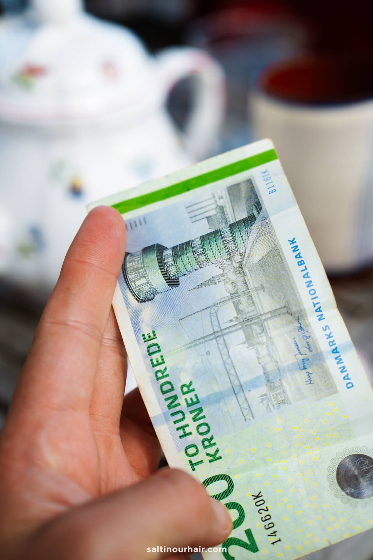 A hand holding a Danish 200 kroner banknote, with a teapot and cup blurred in the background—perfect inspiration as you plan your Denmark itinerary.