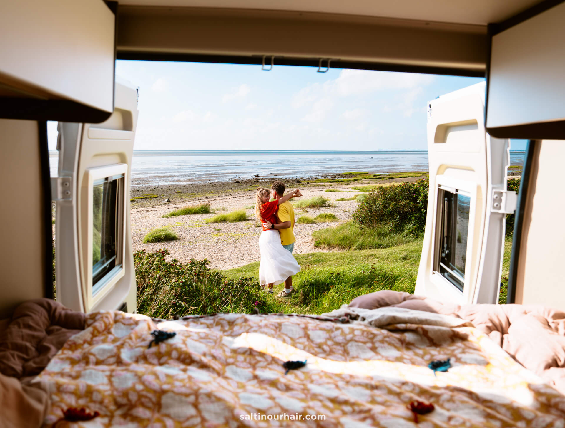 View from inside a camper van looking out at a couple embracing on a grassy area near the beach—an idyllic stop on any Denmark road trip itinerary, with the ocean and sky stretching out in the background.