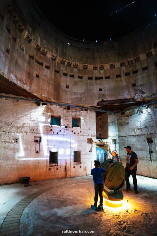 A child and two adults stand inside a large, circular, worn concrete structure at the Tirpitz Museum Denmark, interacting with a lighted display projecting a train image on the wall.