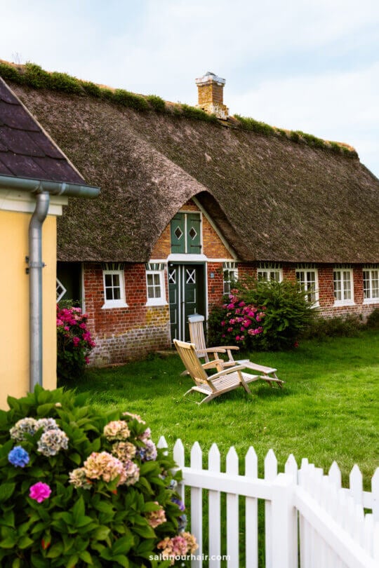 A traditional thatched-roof brick house with a green door in Sønderho, perfect for your Denmark itinerary, is surrounded by flowers and a white picket fence, with two wooden chairs inviting you to relax on the lawn.