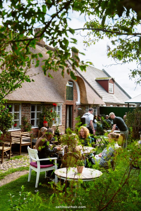 People are sitting at outdoor tables eating and talking in a garden beside a brick house with a thatched roof—a charming stop on any Denmark itinerary—enjoying the sunshine and lively atmosphere.