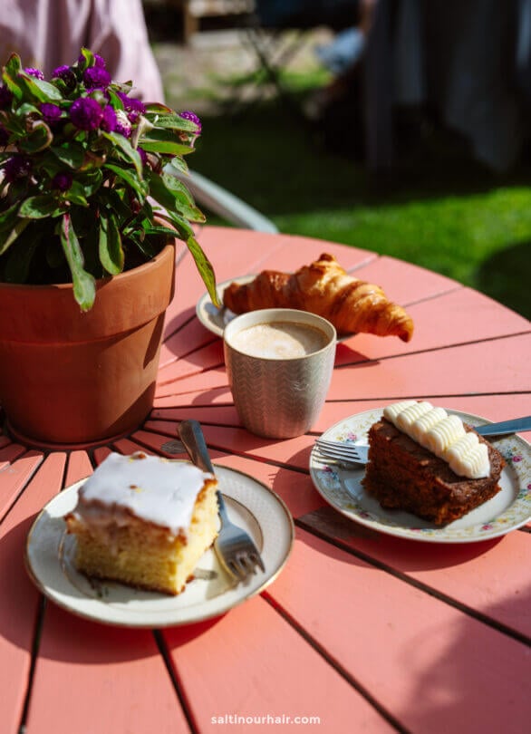 A pink wooden table set outdoors in sunlight, perfect for planning your Denmark itinerary, with a potted plant, a cup of coffee, a croissant, and slices of iced and carrot cake.
