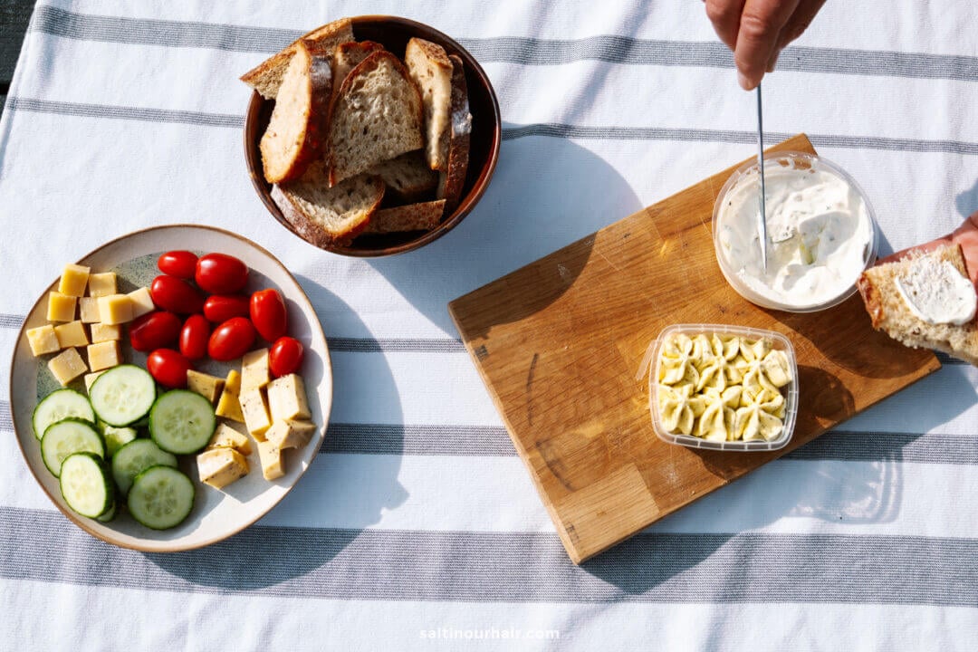 A plate with sliced cucumber, cheese cubes, and cherry tomatoes, a bowl of bread, and a board with cheese spread and chopped cheese on a striped tablecloth—perfect for planning your Denmark itinerary.