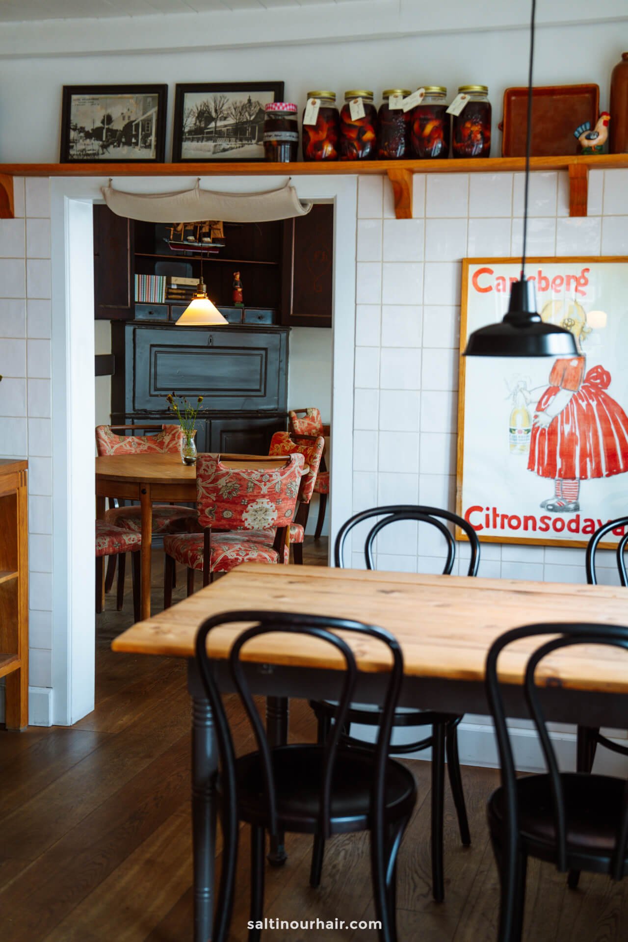 A cozy café interior perfect for planning your Denmark itinerary, with wooden tables and chairs, floral upholstered seating, framed art, and glass jars on a high shelf. A small vase with flowers sits on a table in the background.