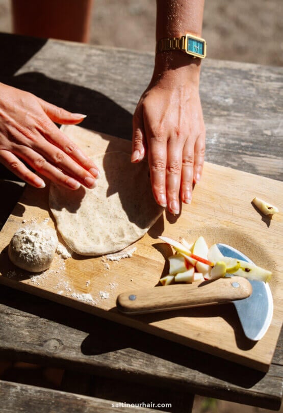Two hands flatten dough on a wooden cutting board next to a ball of dough, sliced apples, a dough cutter, cheese pieces—perfect inspiration for planning recipes into your Denmark itinerary.