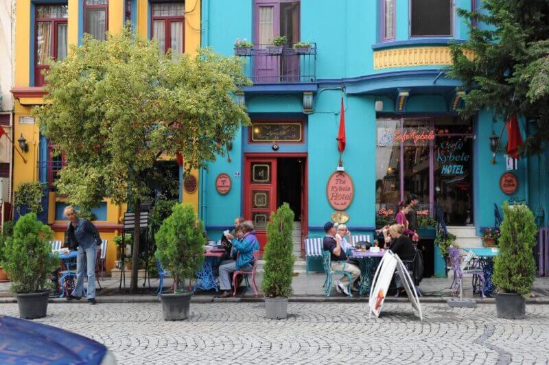 People sit at outdoor tables in front of a colorful, blue-painted hotel and café—one of the charming hotels in Turkey—on a cobblestone street lined with potted plants and trees.