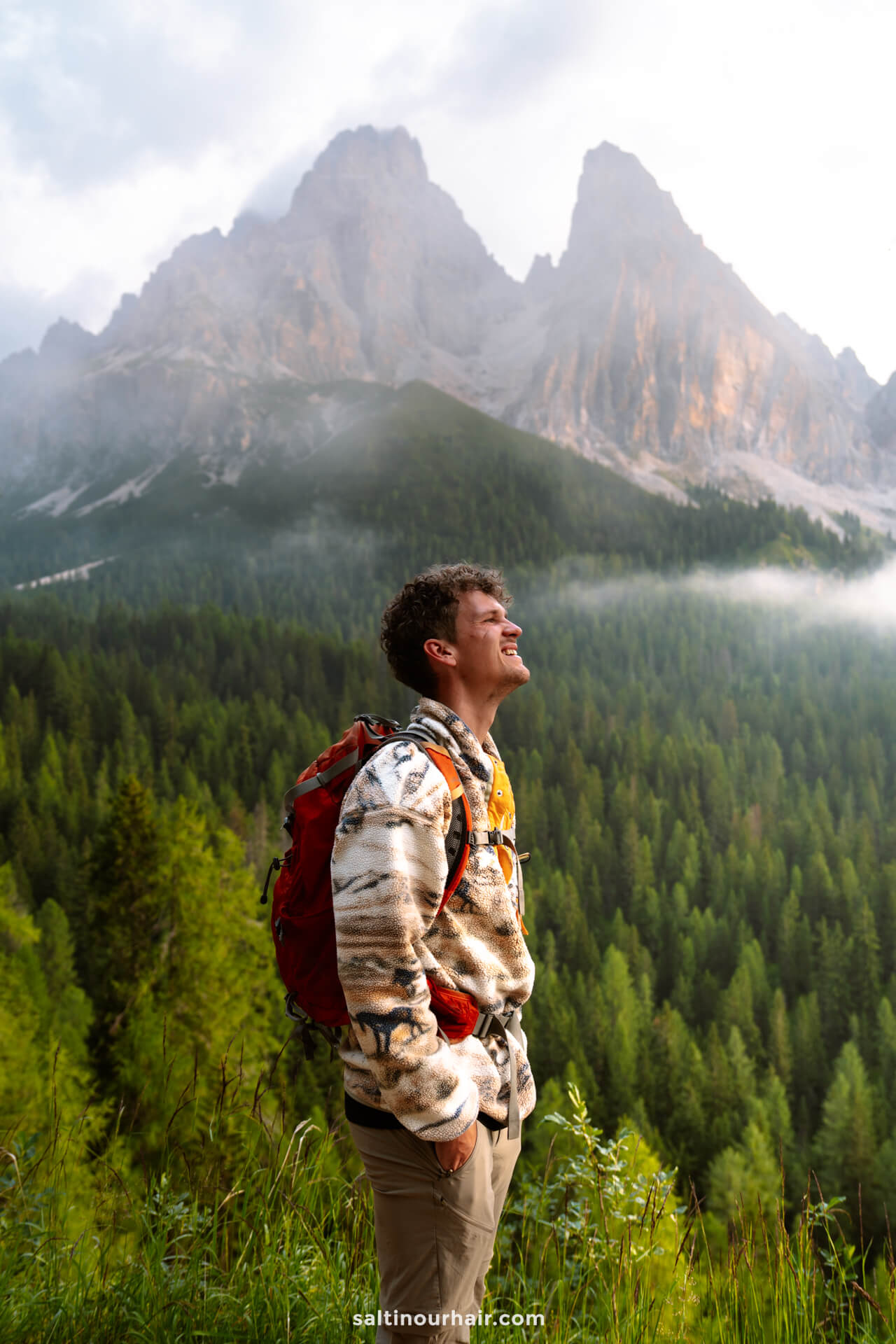 A man wearing a patterned jacket and red backpack stands on a green hillside, looking up toward the misty, forested mountains near Lago di Sorapis, dolomites italy