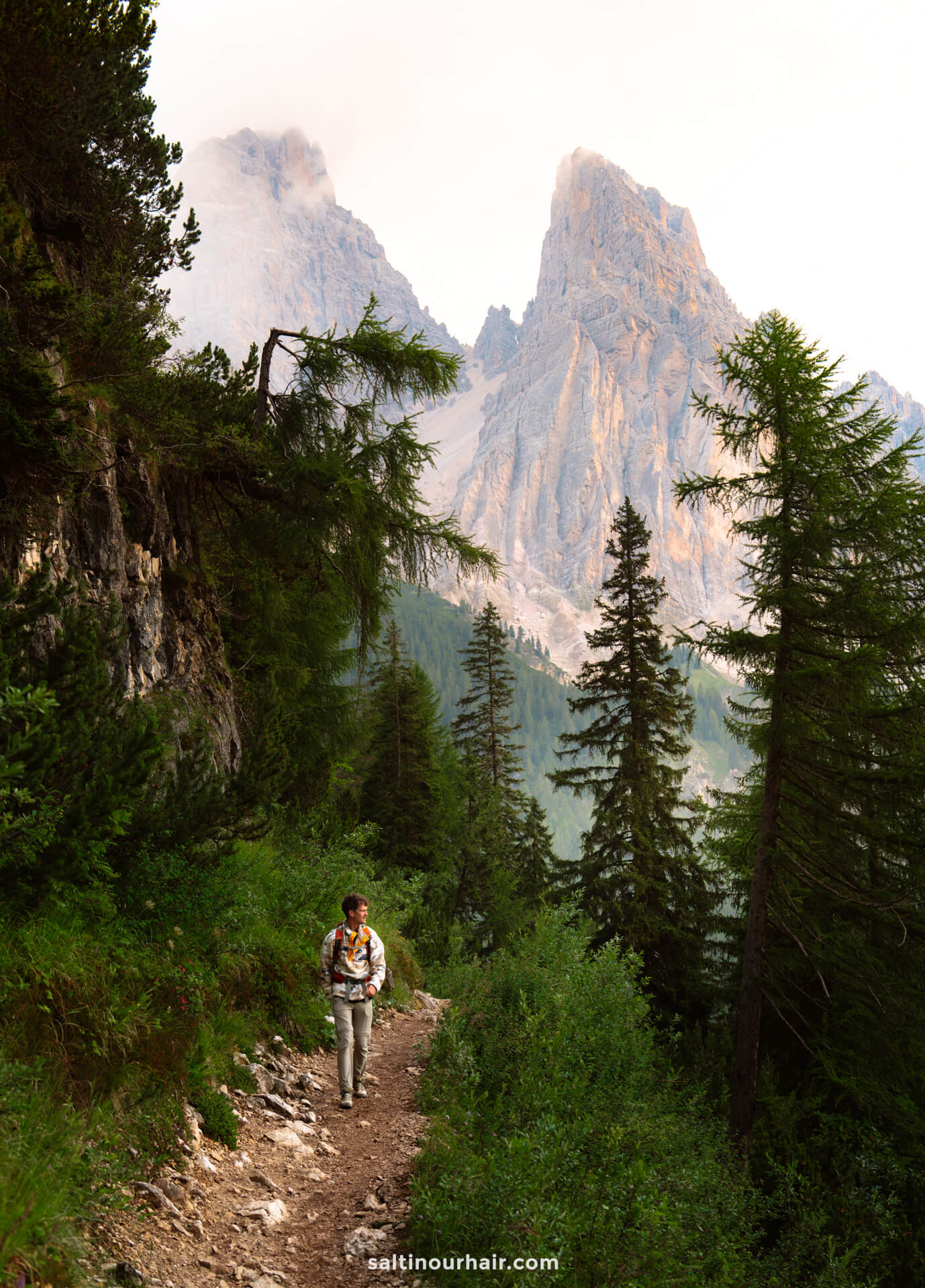 A person enjoys a hike to Lago di Sorapis in the Dolomites, Italy, following a narrow forest trail lined with tall pine trees and rocky mountain peaks in the background.