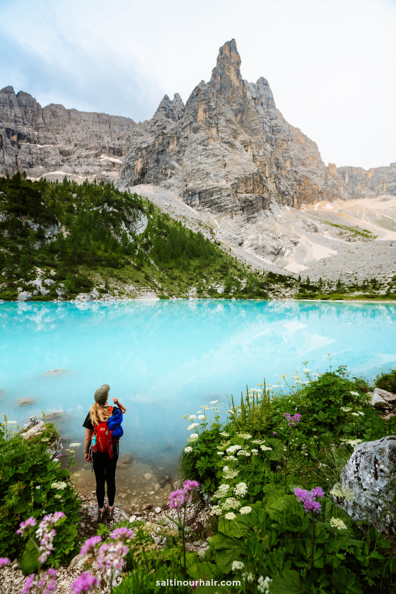 A person with a backpack stands near the edge of turquoise Lago di Sorapis, surrounded by wildflowers, with rocky mountains rising majestically in the background.