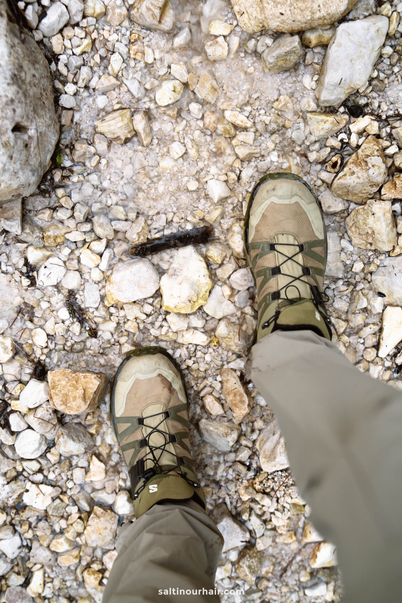 Person wearing tan hiking boots and beige pants standing on a rocky, wet surface near lago di Sorapis, with small stones scattered around their feet.
