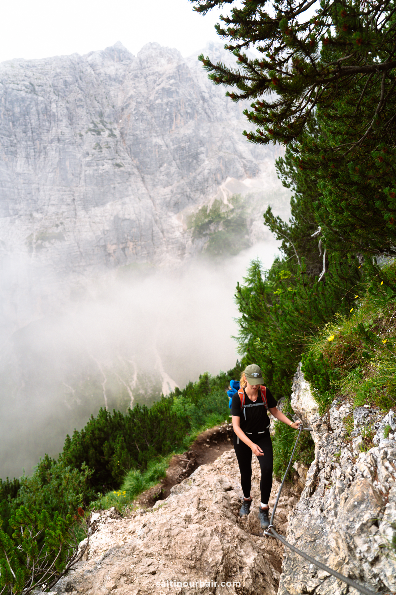 A hiker with a backpack ascends a rocky mountain trail using a rope, surrounded by green trees and misty cliffs near Lago di Sorapis in the Dolomites, Italy.