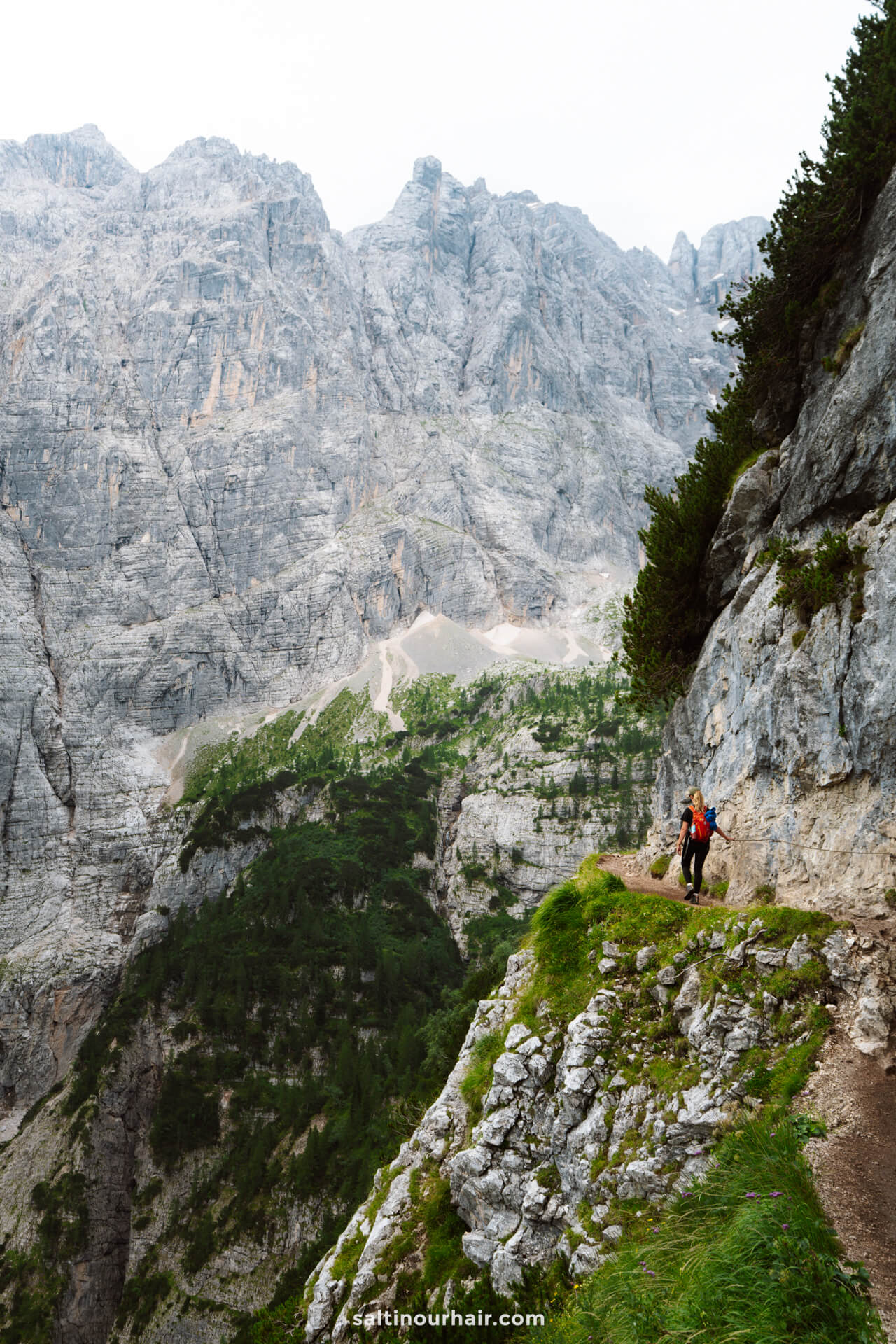 A hiker with a red backpack walks along a narrow mountain trail near Lago di Sorapis, surrounded by steep rocky cliffs and dense green vegetation.