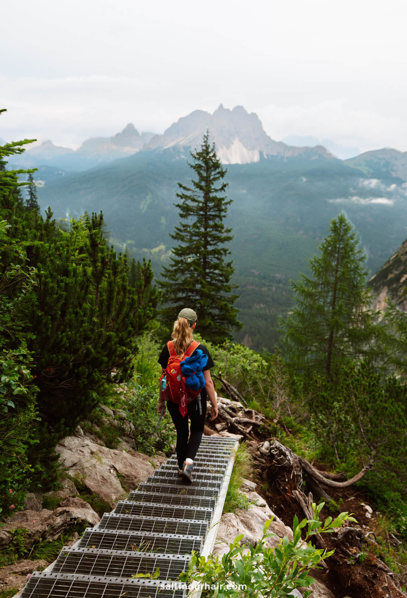 A person with a backpack walks down metal stairs on a mountain trail near Lago di Sorapis, surrounded by green trees, with distant peaks and a cloudy sky in the background.