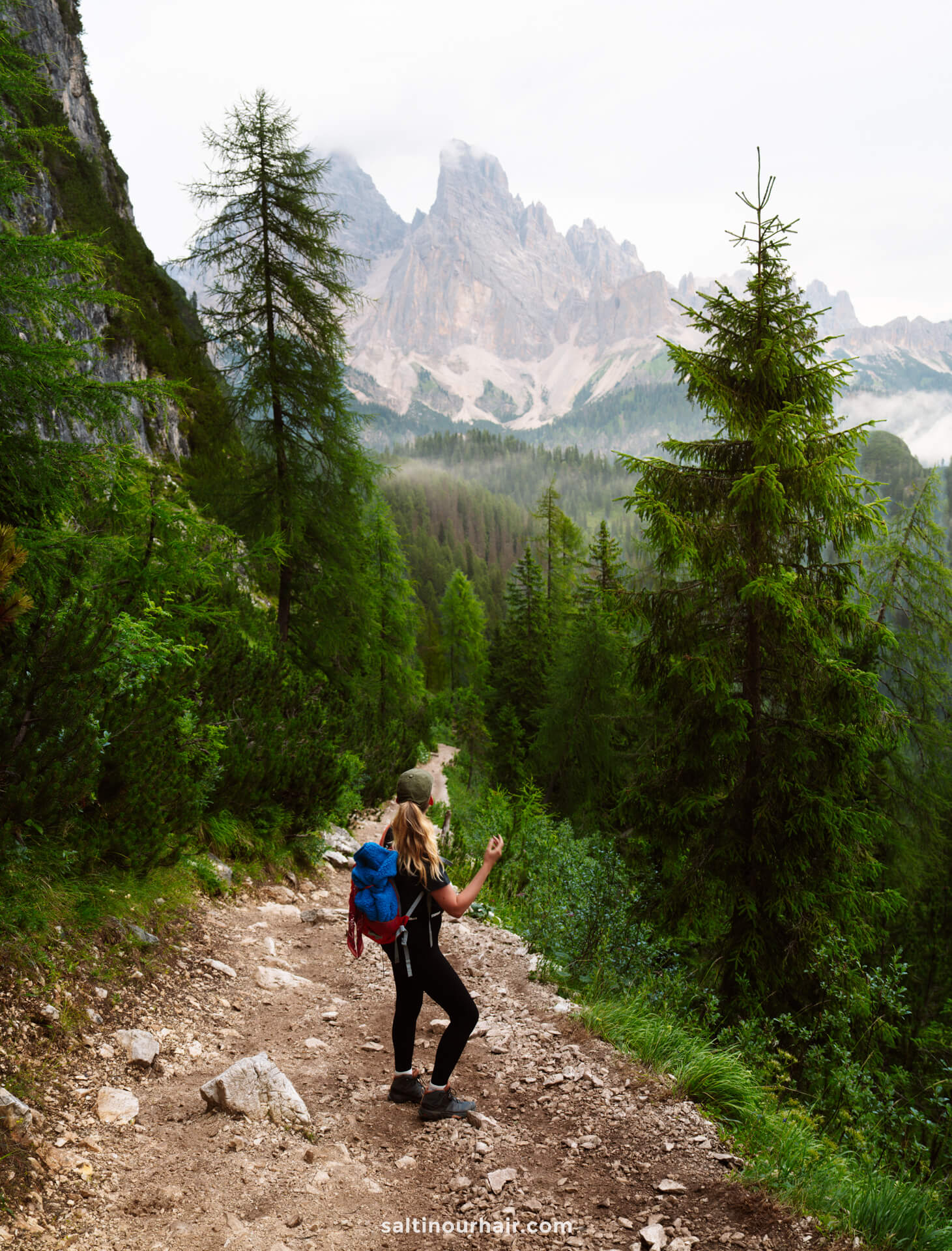 A person with a blue backpack hikes on a rocky mountain trail near Lago di Sorapis, surrounded by green trees, with tall peaks visible in the distance under a cloudy sky.
