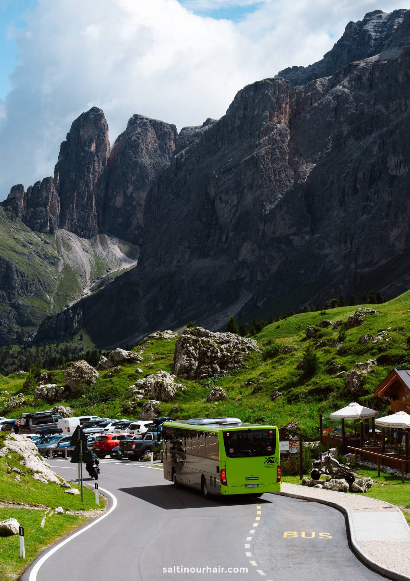A green bus drives on a winding road near a parking lot, with rocky mountains and grassy hills in the background, not far from the picturesque Lago di Sorapis.