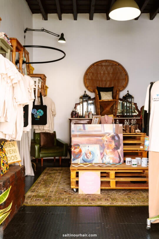 A small boutique in San Crist&oacute;bal de La Laguna tenerife displays clothing, tote bags, art prints, mugs, and decorative lanterns, with a large wicker chair and various items arranged on wooden pallets and shelves.
