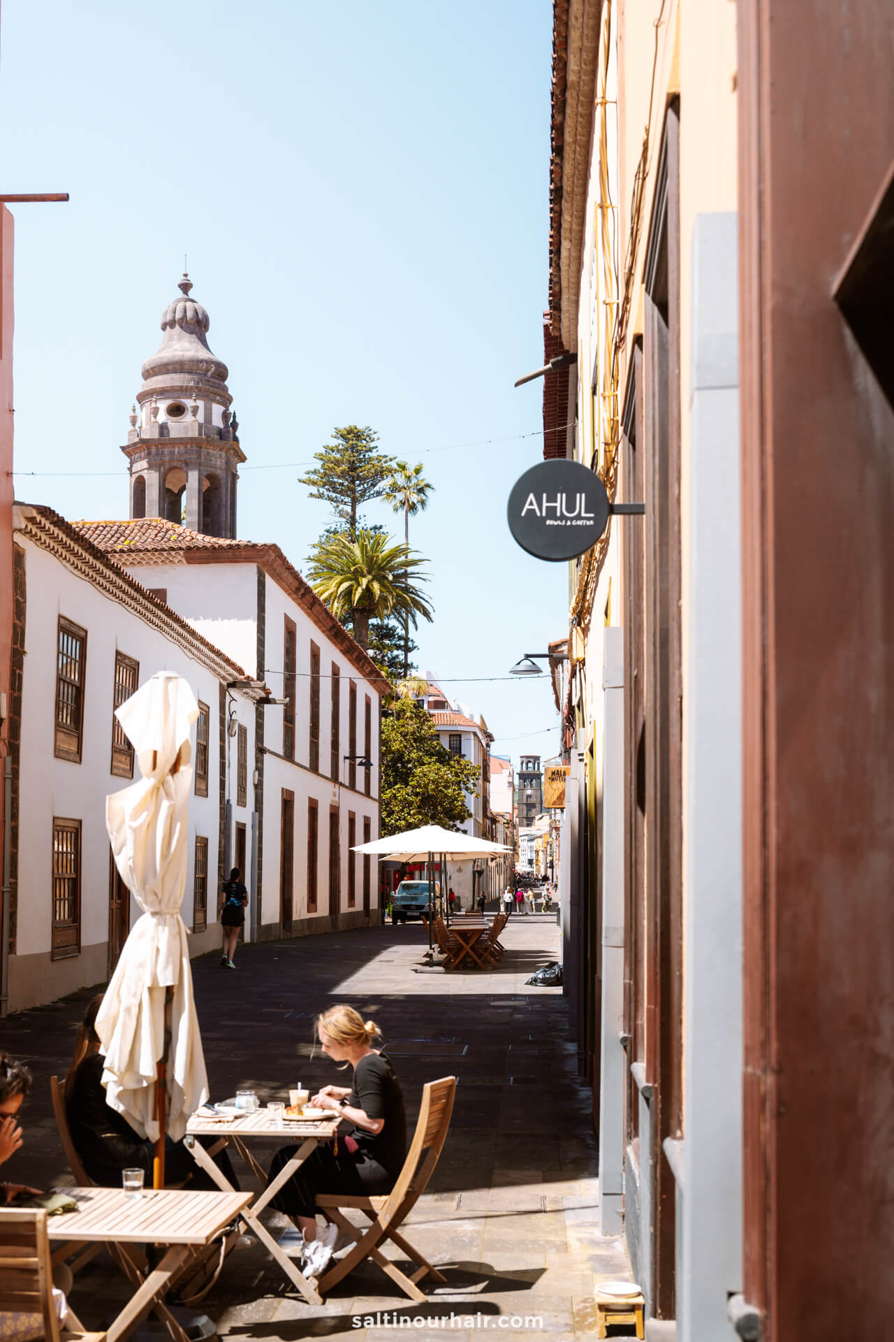 A narrow street in San Crist&oacute;bal de La Laguna tenerife with outdoor cafe seating, people dining, white buildings, and La Laguna Cathedral in the  background.