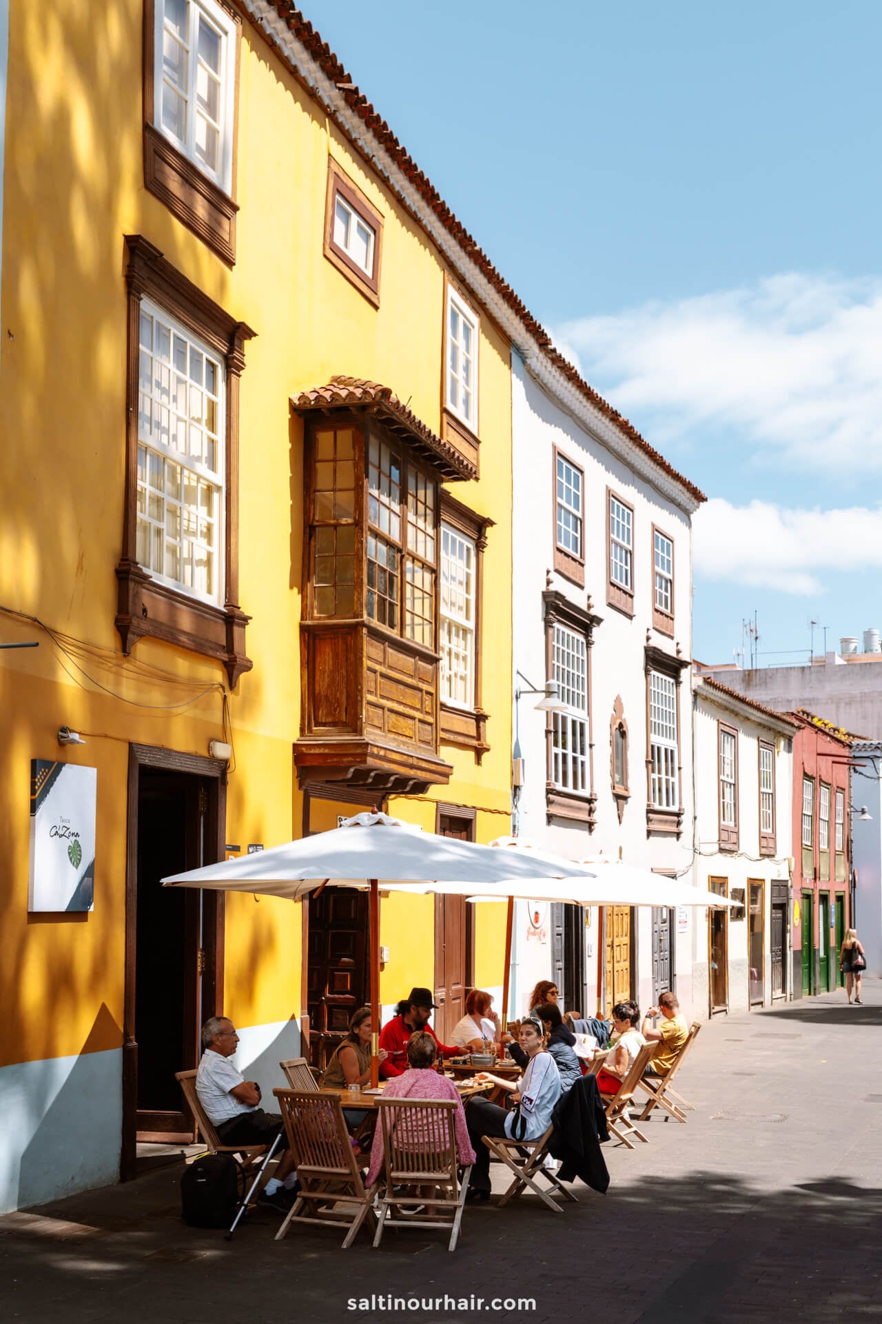 People sit at tables under umbrellas outside a cafe on a sunny street in San Crist&oacute;bal de La Laguna tenerife, lined with colorful, traditional European-style buildings.