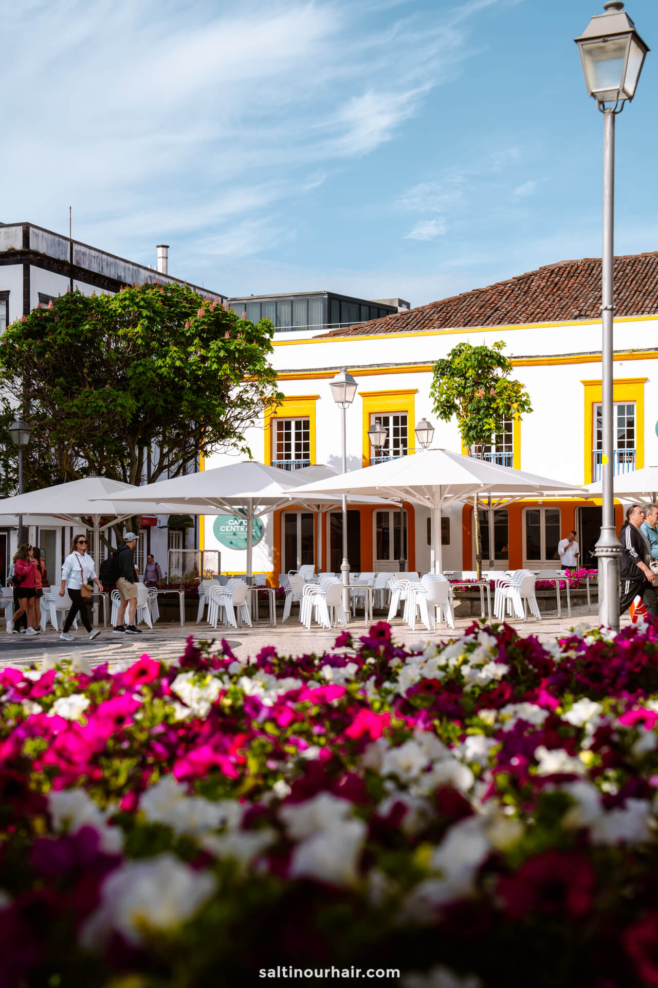 Outdoor caf&eacute; with white umbrellas and chairs in ponta delgada azores, surrounded by people and vibrant pink and white flowers, set against a white and yellow building