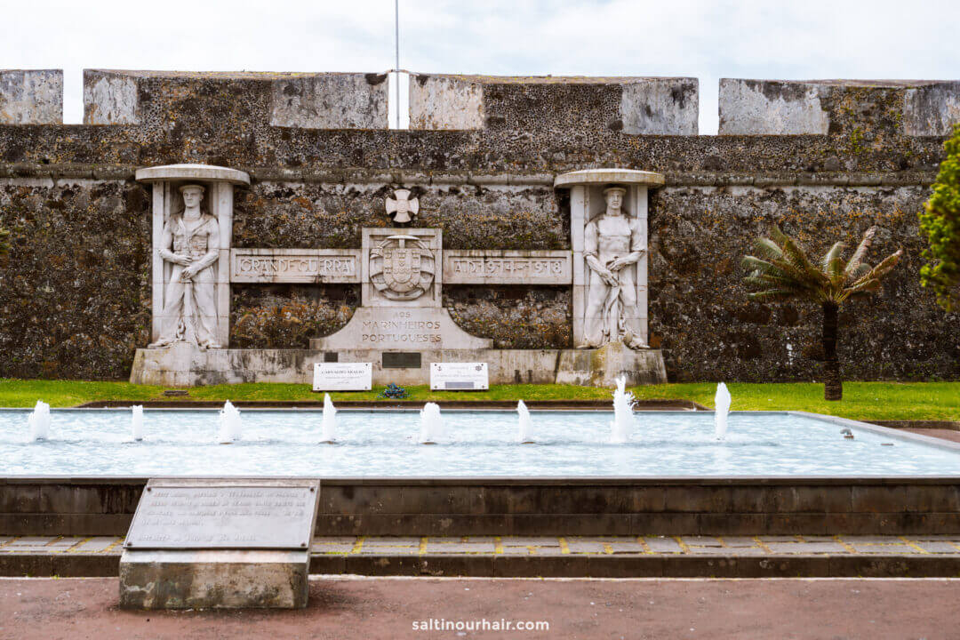 A stone monument with two statues stands in front of a fortress wall, with a rectangular fountain and plaque in the foreground