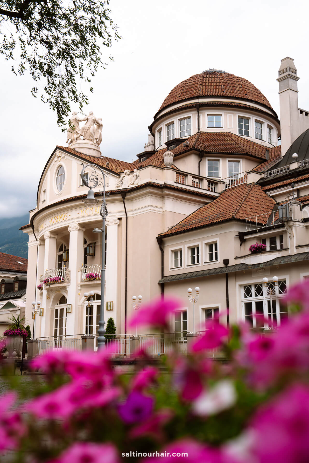 A historic building in Merano, Italy with a domed roof and ornate facade, seen behind pink flowers in the foreground.