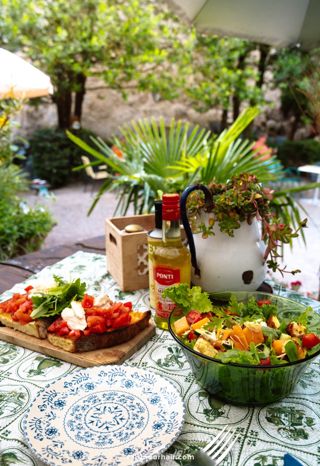 A table outdoors in Merano Italy displays a salad, bruschetta with tomato and cheese, a bottle of olive oil, and a potted plant, with a picturesque garden visible in the background.