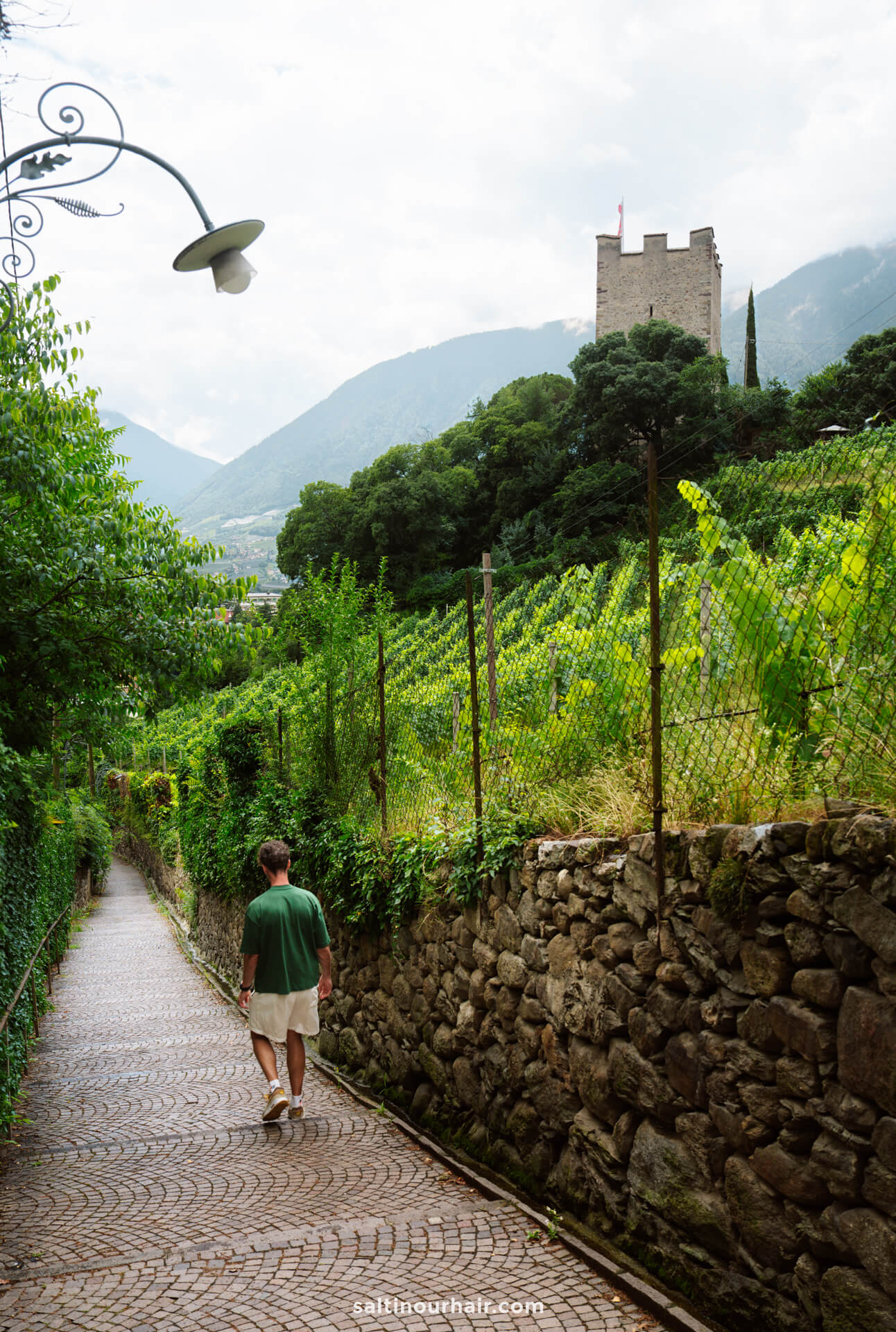 A person walks along a narrow cobblestone path lined with stone walls and greenery in Merano, Dolomites Italy, with a tower and mountains visible in the background.
