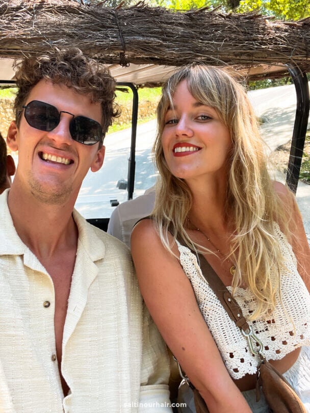 Two people sitting in an open vehicle on a sunny day, smiling at the camera. Both are wearing light, summery clothing and sunglasses, enjoying the farm to table experience in Tuscany, Italy.