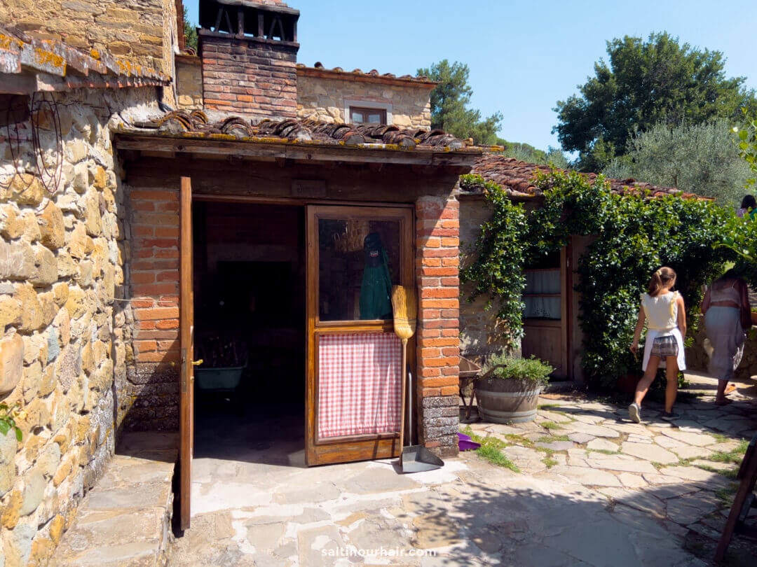 Stone and brick building with a wooden door, red checkered curtain, and broom outside; two people walk away on a sunny day, evoking the charm of a Tuscany farm in Italy.