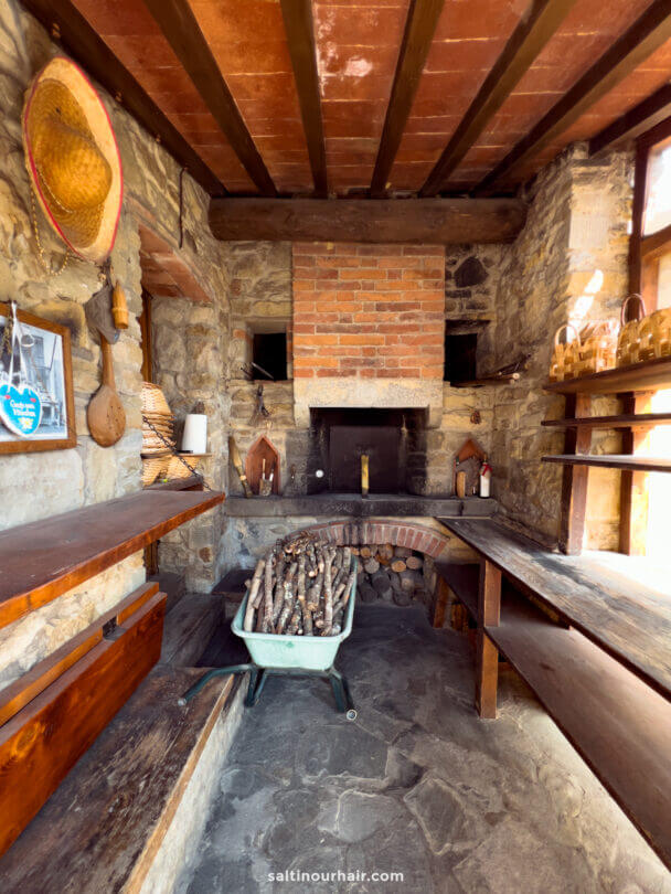 A rustic stone-walled room on a Tuscany farm, featuring a fireplace, wooden shelves, a wheelbarrow filled with firewood, and various tools hanging on the walls.