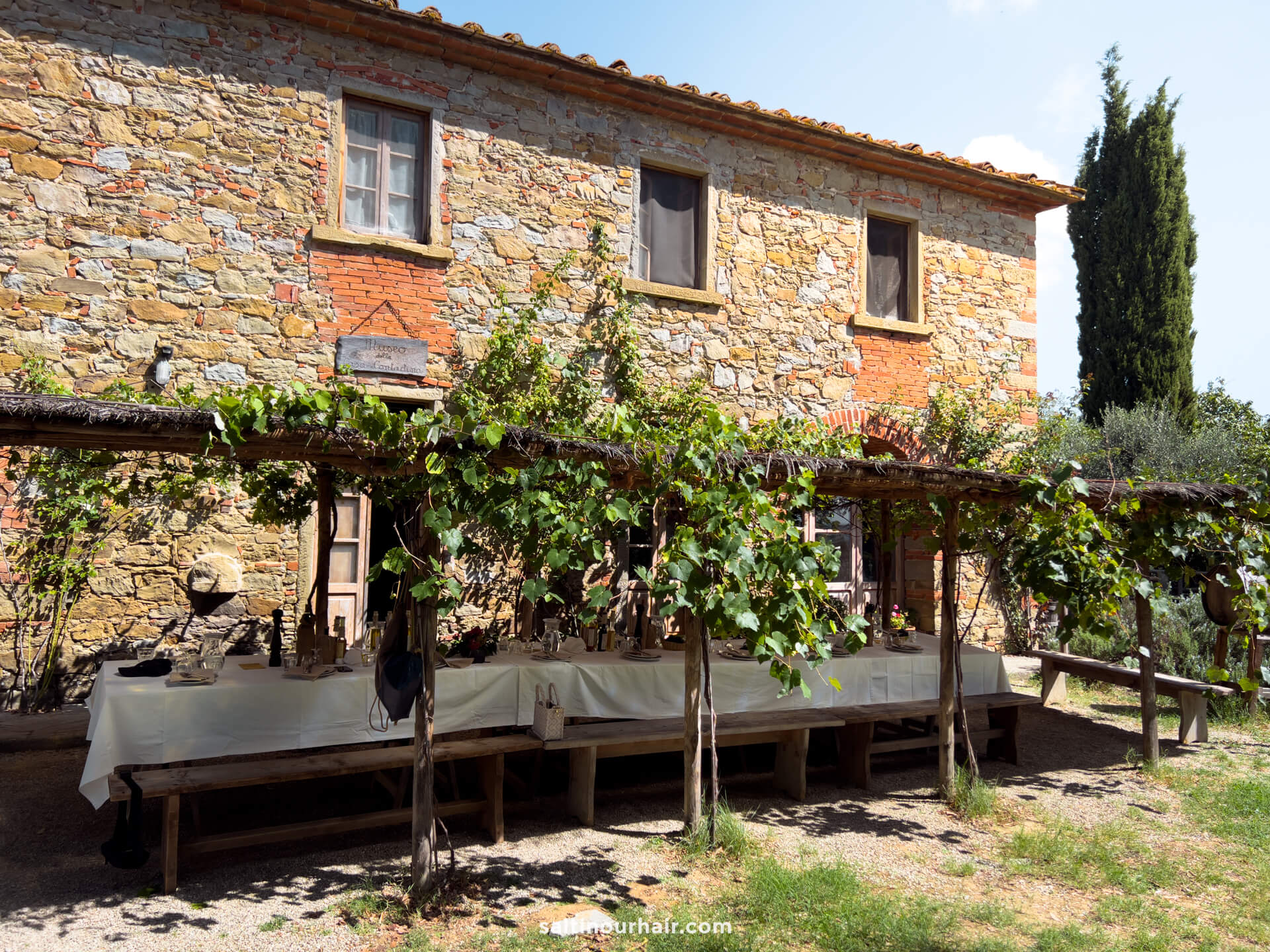 A long outdoor dining table is set under a grapevine-covered pergola Fattoria la Vialla Tuscany Italy, in front of a rustic stone house.