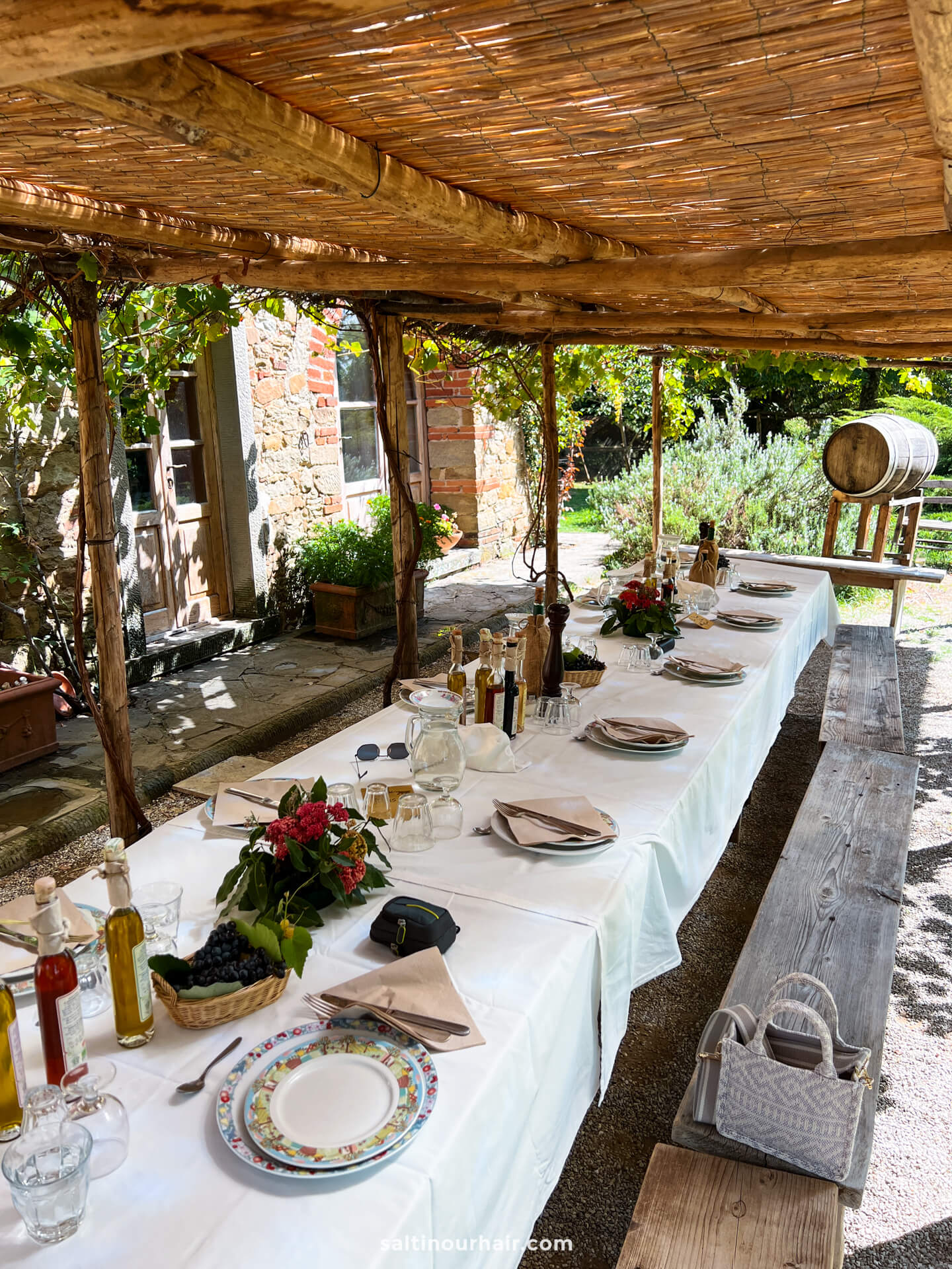 A long outdoor table is set for a meal under a wooden pergola with a white tablecloth, plates, glassware, and bottles&mdash;evoking the rustic charm of a farm in Tuscany Italy beside a stone building and lush greenery.