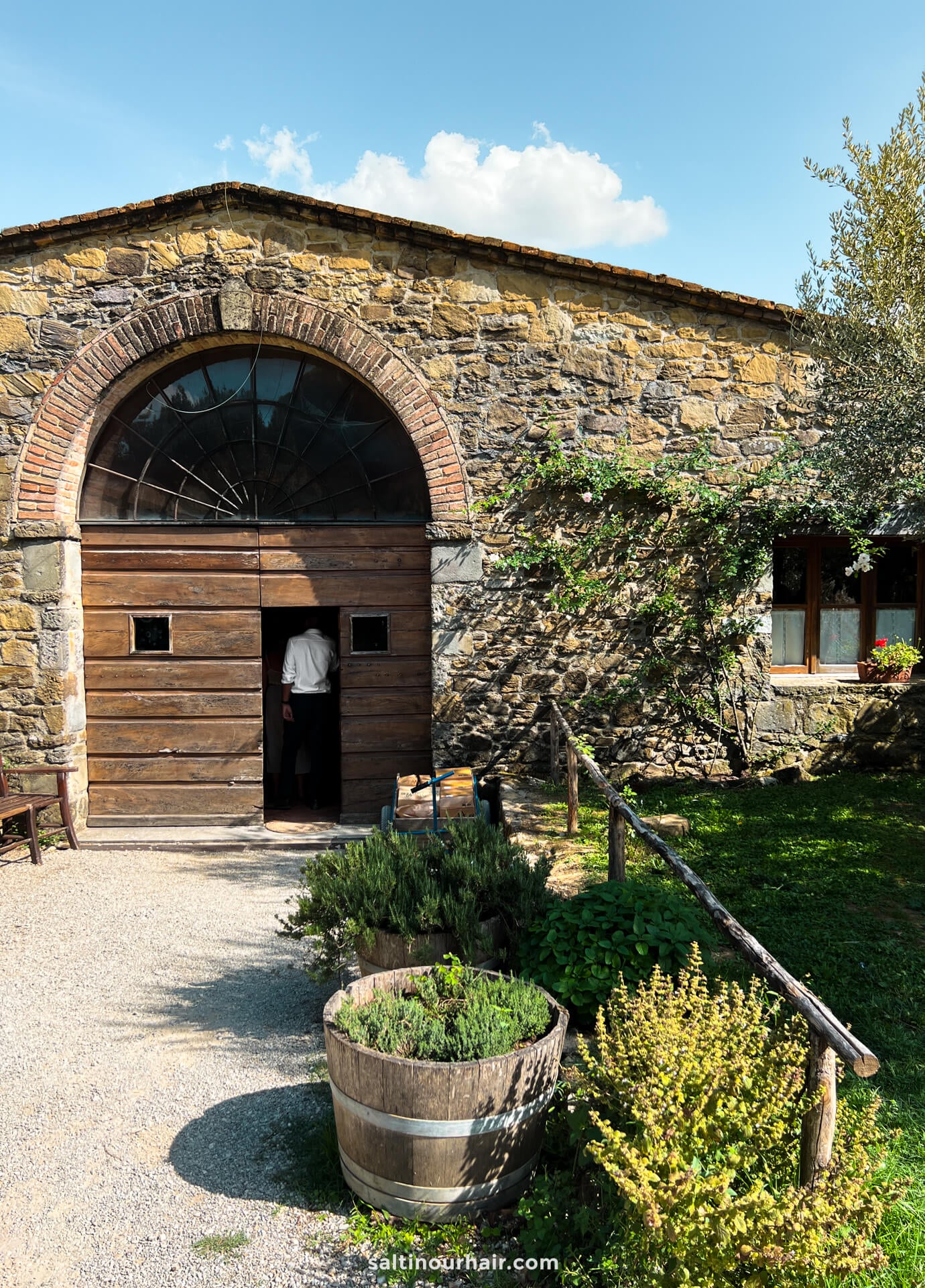 A person enters a rustic stone building with a large wooden door, reminiscent of a farm in Tuscany italy, surrounded by plants and a wooden barrel planter.