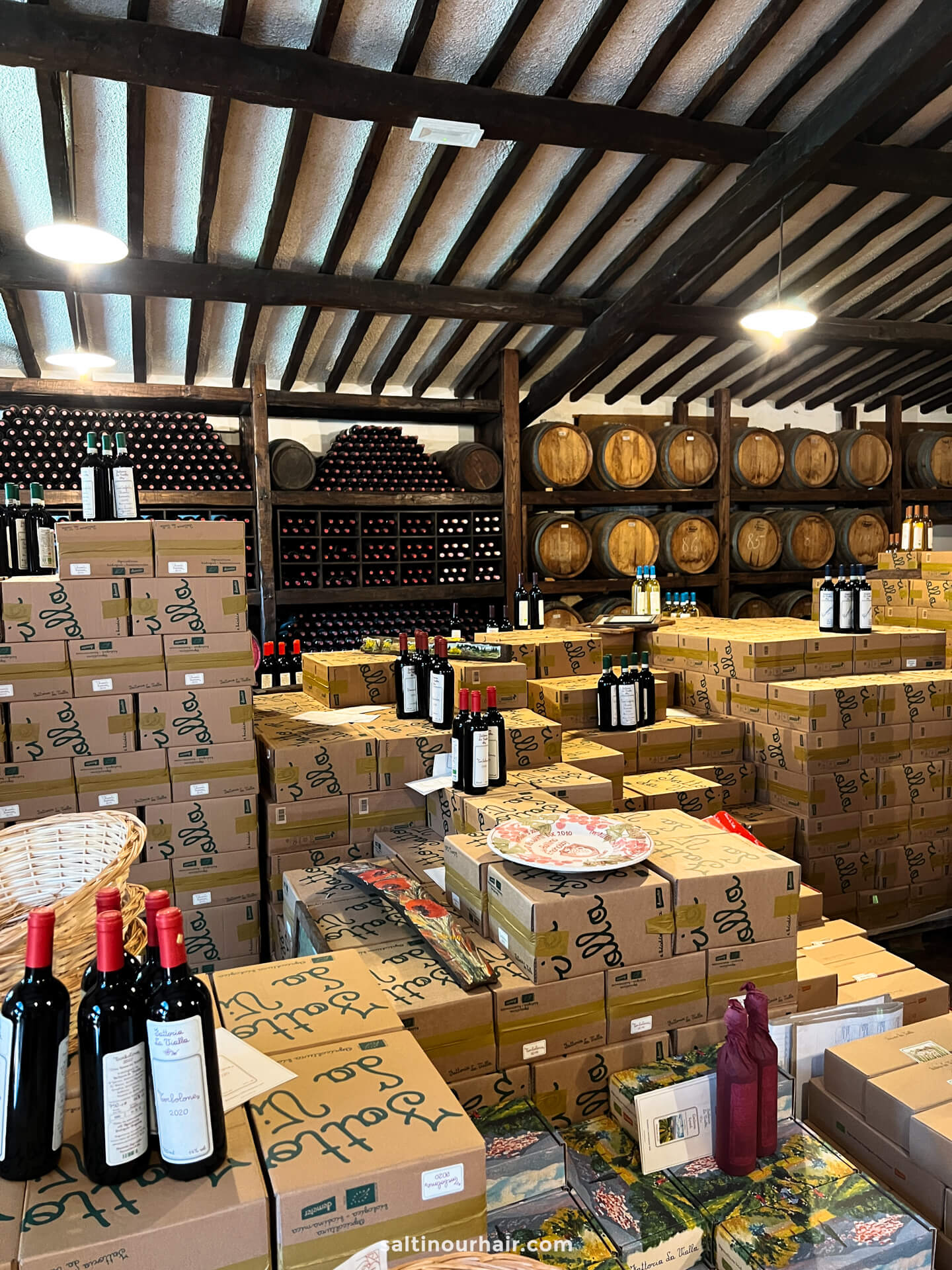 Stacks of cardboard boxes and bottles of wine are arranged at the cellar of Fattoria la Vialla tuscany italy, with wooden barrels on shelves and wine bottles displayed in the background.