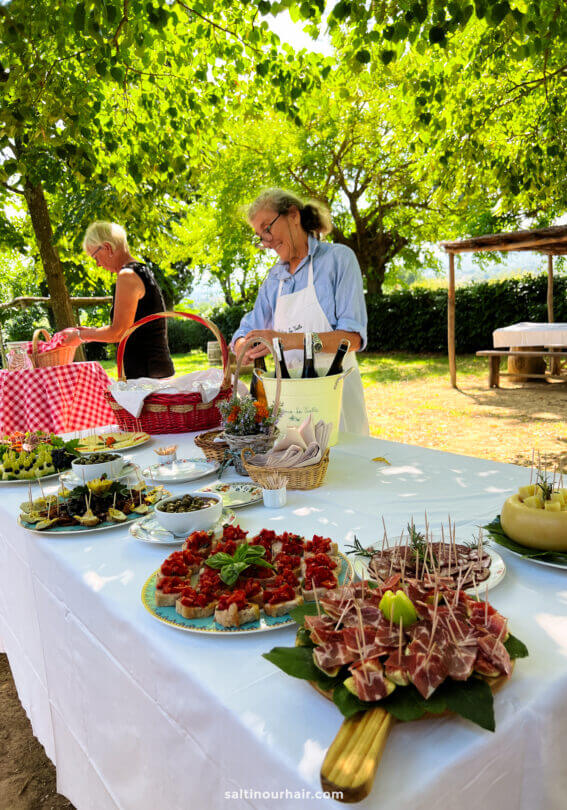 Two women prepare and arrange appetizers on outdoor tables covered with white and checkered cloths, surrounded by greenery and shaded by trees on a charming Tuscany farm.
