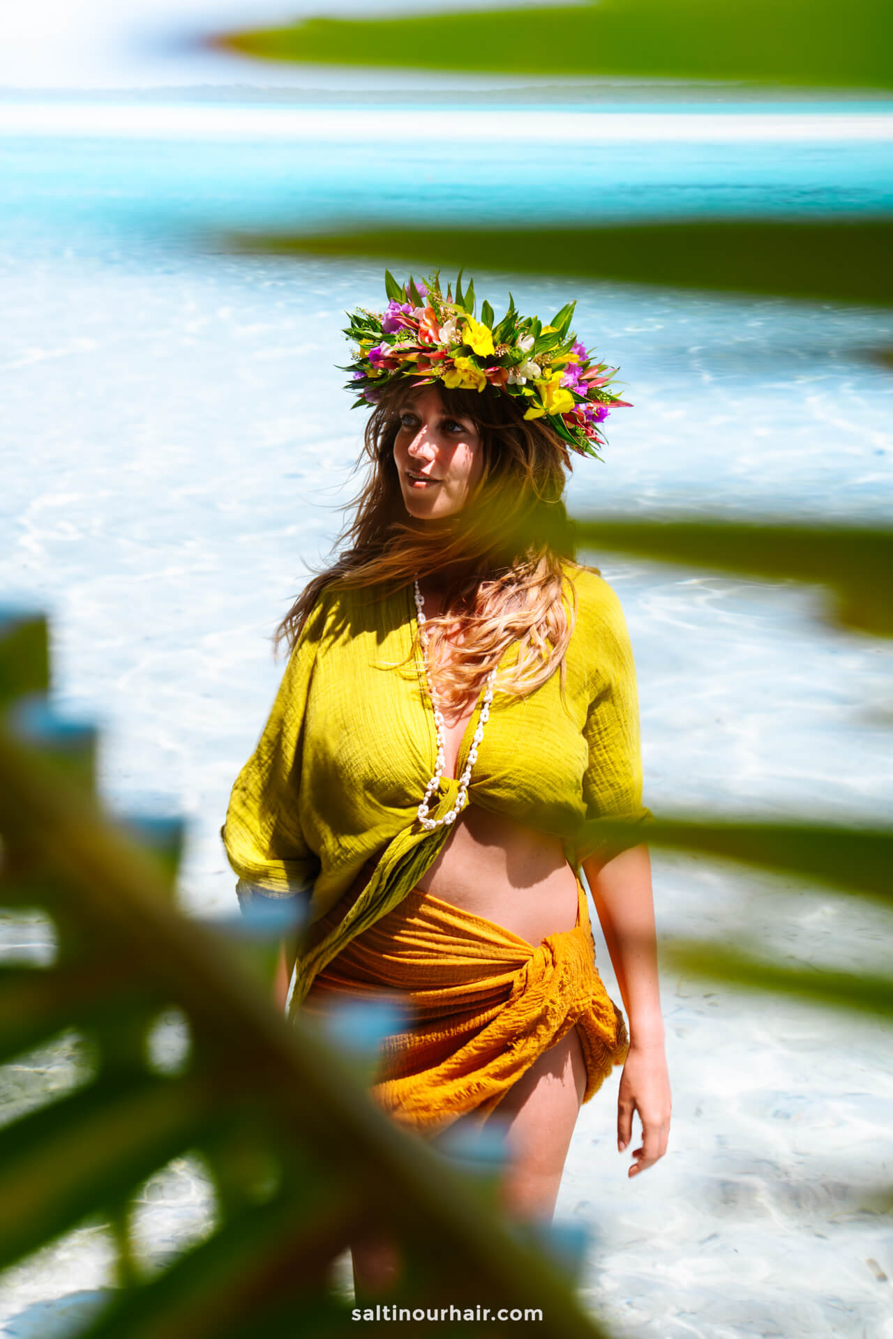 A woman wearing a yellow-green top, orange wrap skirt, and a floral crown stands in shallow, clear water on Aitutaki island, partially obscured by palm leaves.