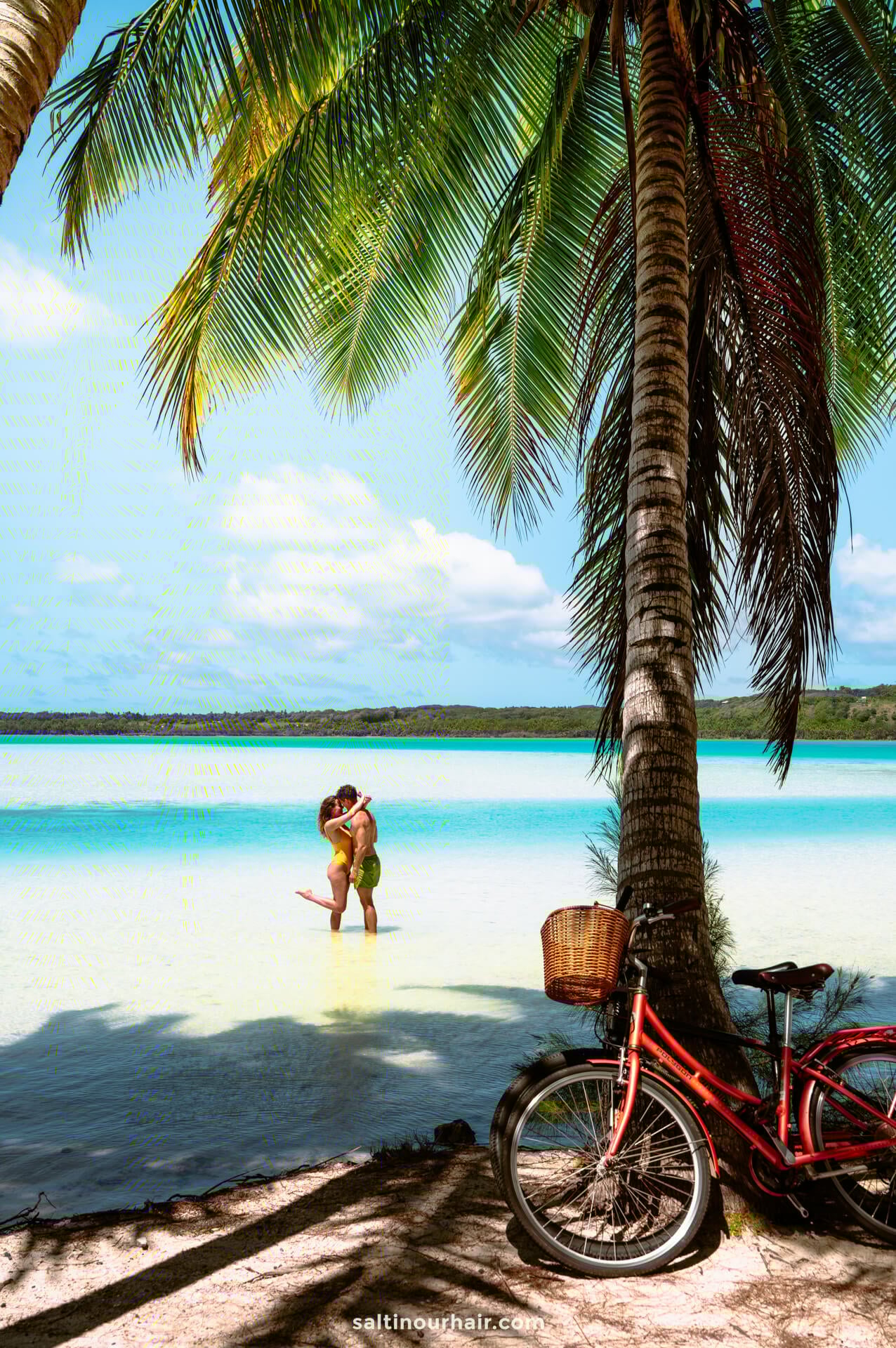 A couple stands embracing in shallow turquoise water under palm trees on Aitutaki island, with a red bicycle and basket resting on the sandy shore in the foreground.