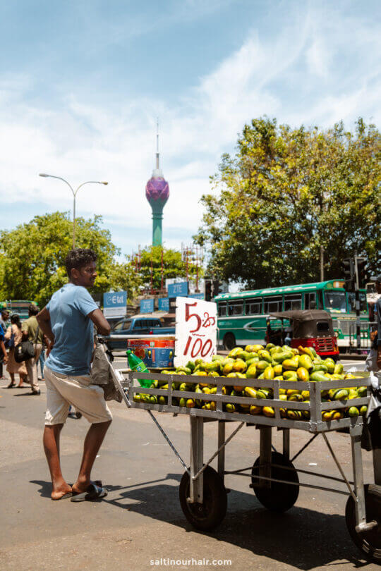 A man stands next to a street cart selling mangoes with a sign reading 5 for 100, set against bustling buses and the iconic Lotus Tower&mdash;one of the things to do in Colombo.