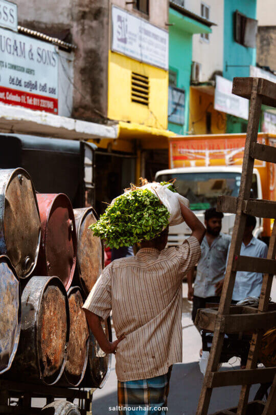 A man carries a bundle of green leafy vegetables on his head while walking down a busy street in Pettah Market Colombo lined with barrels and shops.