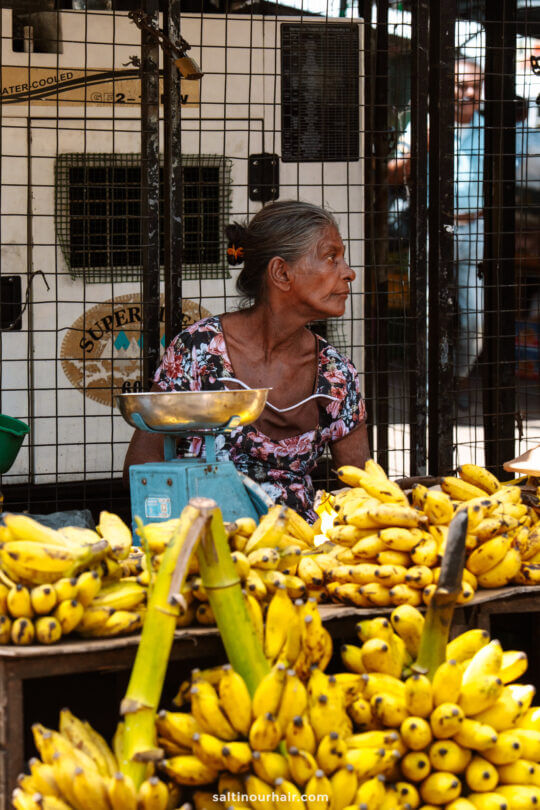 An older woman sits behind a display of bananas at a market stall in Colombo, with a weighing scale in front of her