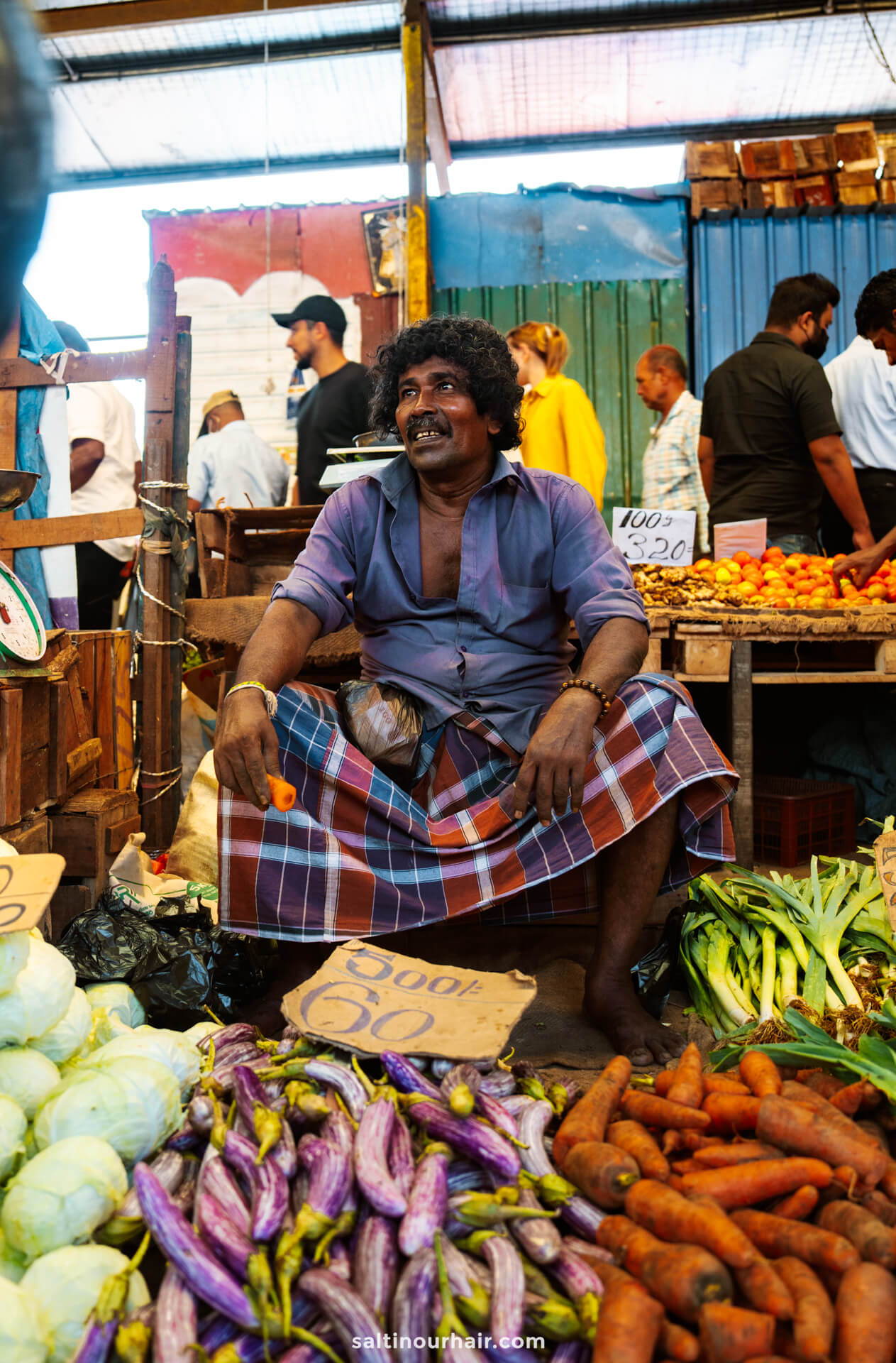 A vendor sits among his vegetables, including eggplants and carrots, at an outdoor market&mdash;a vibrant scene that