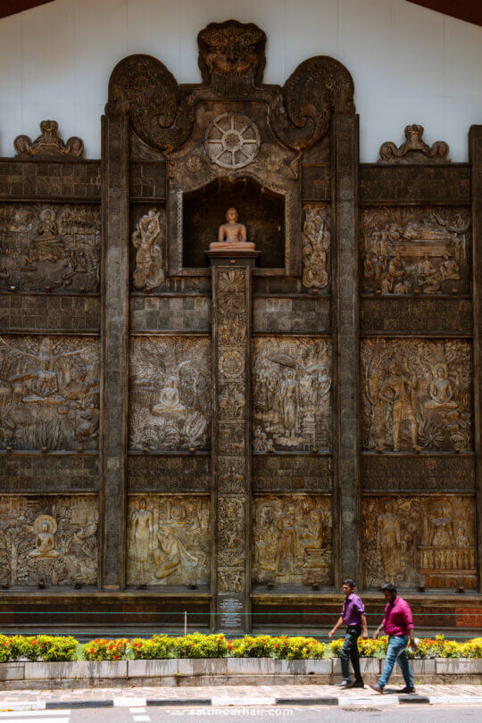Two people walk in front of a large intricately carved stone wall featuring religious and historical reliefs, with a Buddha statue in a niche above the central column.