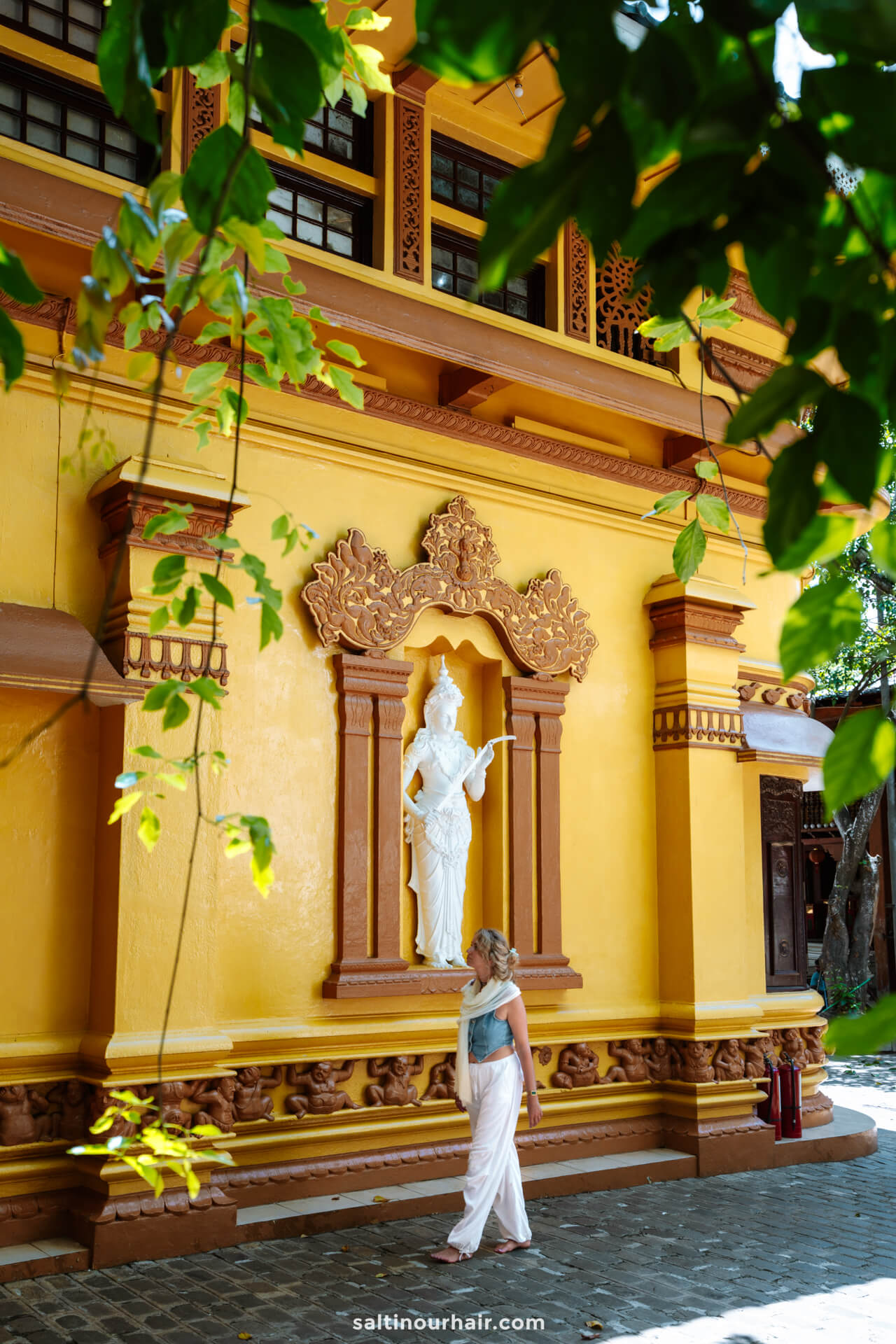 A woman walks past a yellow temple wall featuring a white statue set in an ornate niche, with green leaves framing the scene&mdash;a hidden gem among the many things to do in Colombo.